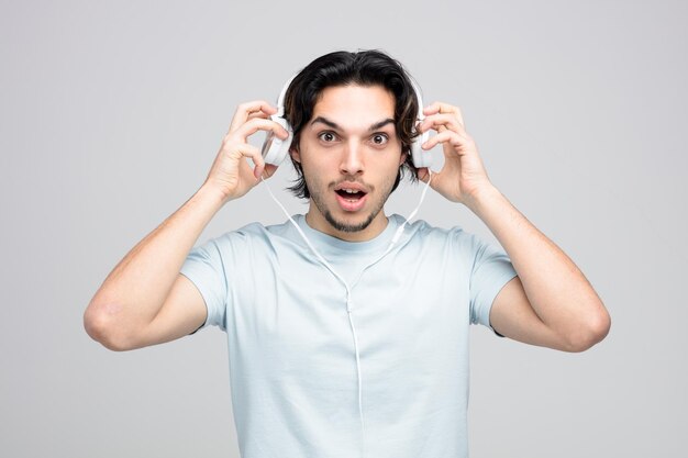 Impresionado joven guapo mirando a la cámara quitándose los auriculares aislado sobre fondo blanco.