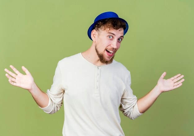 Impresionado joven guapo partido eslavo con gorro de fiesta mirando a la cámara mostrando las manos vacías aisladas sobre fondo verde oliva