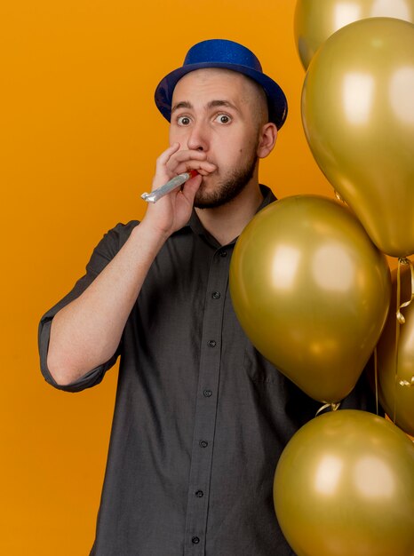 Impresionado joven guapo partido eslavo con sombrero de fiesta sosteniendo globos soplador de fiesta mirando a cámara aislada sobre fondo naranja