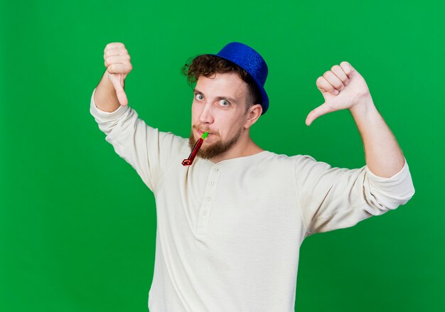 Impresionado joven guapo partido eslavo con sombrero de fiesta sosteniendo el soplador de fiesta en la boca mirando a la cámara mostrando los pulgares hacia abajo aislado sobre fondo verde