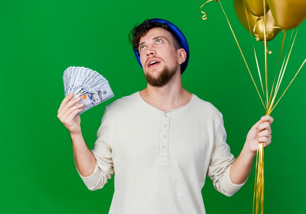 Impresionado joven guapo partido eslavo vistiendo gorro de fiesta sosteniendo globos y dinero mirando hacia arriba aislado sobre fondo verde