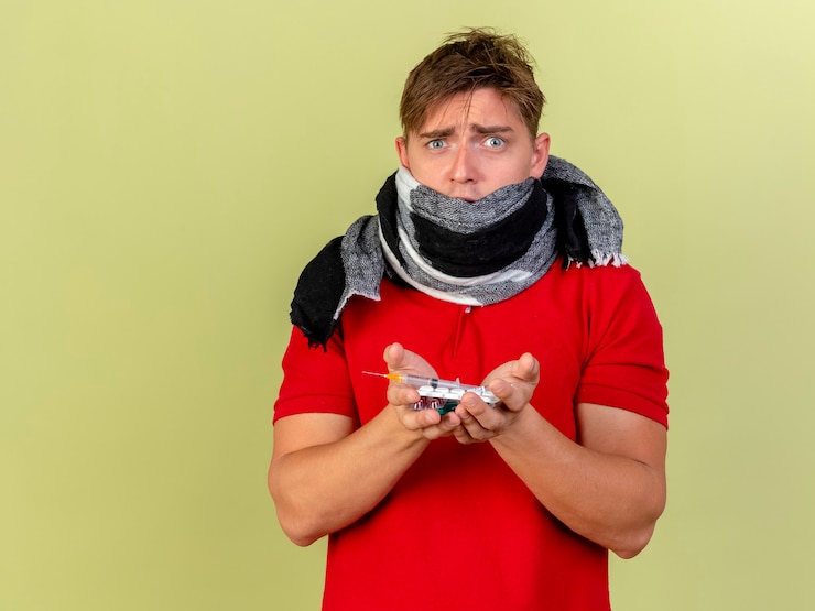 Foto gratuito impresionado joven guapo rubio enfermo con bufanda sosteniendo píldoras médicas y una jeringa aislada en la pared verde oliva con espacio de copia