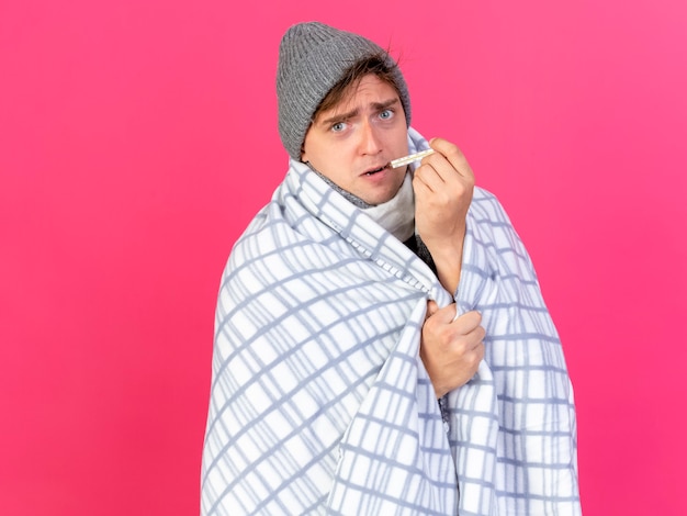 Impresionado joven guapo rubio enfermo con gorro de invierno y bufanda envuelto en plaid poniendo el termómetro en la boca mirando al frente aislado en la pared rosa