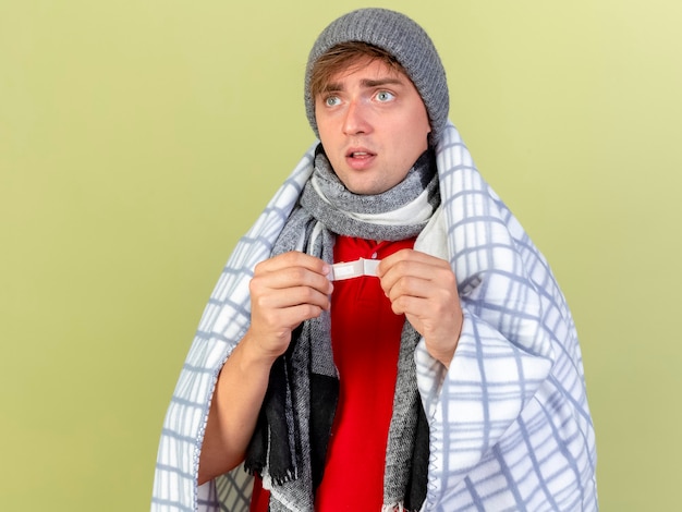 Impresionado joven guapo rubio enfermo con gorro de invierno y bufanda envuelto en plaid sosteniendo yeso mirando al lado aislado sobre fondo verde oliva con espacio de copia