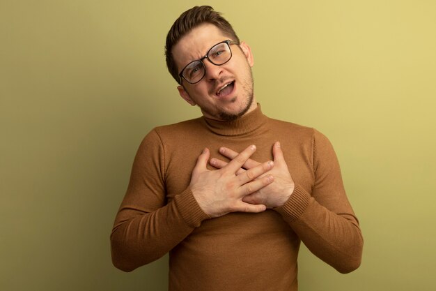 Impresionado joven guapo rubio con gafas mirando poniendo las manos en el pecho aislado en la pared verde oliva con espacio de copia