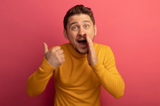 Impresionado joven guapo rubio mirando manteniendo la mano cerca de la boca apuntando detrás susurrando aislado en la pared rosa