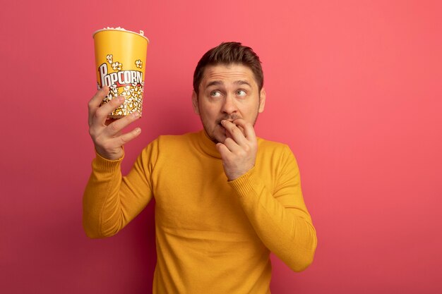 Impresionado joven guapo rubio sosteniendo un cubo de palomitas de maíz y comiendo palomitas de maíz mirando al lado aislado en la pared rosa con espacio de copia