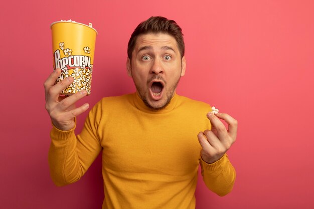 Impresionado joven guapo rubio sosteniendo un cubo de palomitas de maíz y una pieza de palomitas de maíz