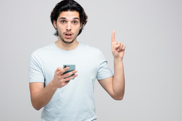 Impresionado joven guapo sosteniendo teléfono móvil mirando a la cámara apuntando hacia arriba aislado sobre fondo blanco.