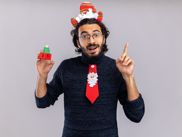 Impresionado joven guapo vistiendo corbata de Navidad con aro de pelo sosteniendo puntos de juguete de Navidad en arriba aislado en la pared blanca