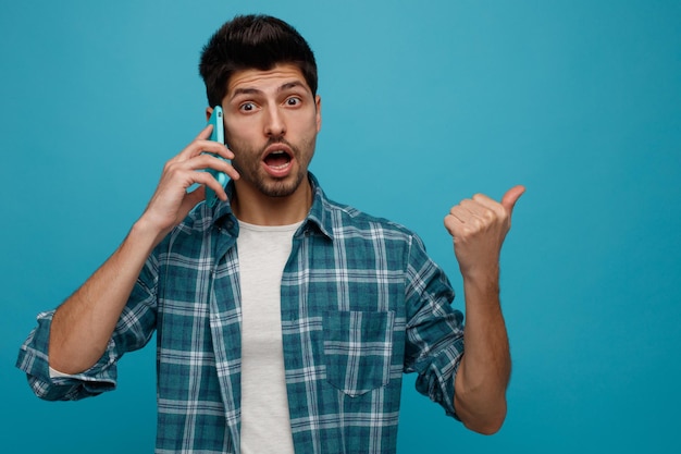 Impresionado joven hablando por teléfono mirando a la cámara apuntando al lado aislado sobre fondo azul.