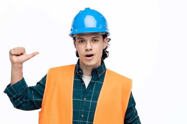 Impresionado joven ingeniero masculino usando casco de seguridad y chaleco de seguridad mirando a la cámara apuntándose a sí mismo aislado en fondo blanco