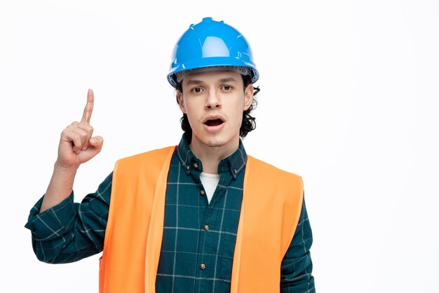 Impresionado joven ingeniero usando casco de seguridad y chaleco de seguridad mirando a la cámara apuntando con el dedo hacia arriba aislado en fondo blanco