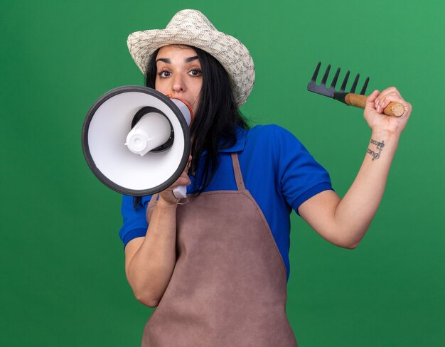 Impresionado joven jardinero caucásico mujer vestida con uniforme y sombrero levantando rastrillo hablando por altavoz