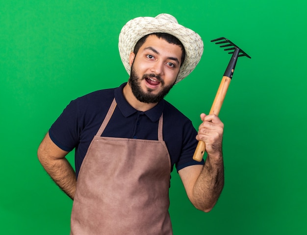 Impresionado joven jardinero varón caucásico vistiendo sombrero de jardinería sosteniendo rastrillo aislado en la pared verde con espacio de copia