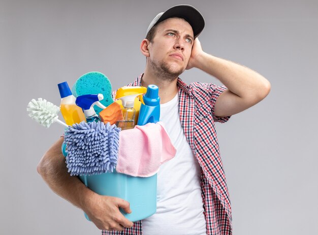 Impresionado joven limpiador con gorra sosteniendo el balde con herramientas de limpieza poniendo la mano en la mejilla aislado sobre fondo blanco.