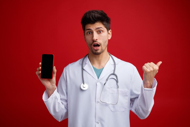 Impresionado joven médico masculino con uniforme médico y estetoscopio alrededor del cuello mirando a la cámara que muestra el teléfono móvil apuntando al lado aislado en el fondo rojo