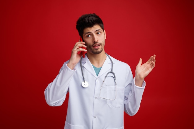 Impresionado joven médico masculino con uniforme médico y estetoscopio alrededor de su cuello hablando por teléfono mostrando la mano vacía mirando al lado aislado en el fondo rojo