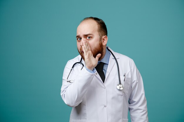 Impresionado joven médico masculino usando abrigo médico y estetoscopio alrededor de su cuello manteniendo la mano cerca de la boca mirando a la cámara susurrando aislado sobre fondo azul.