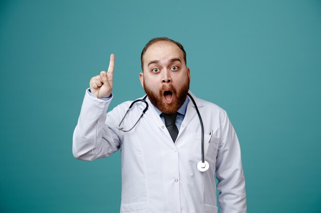 Impresionado joven médico masculino usando abrigo médico y estetoscopio alrededor de su cuello mirando a la cámara apuntando hacia arriba aislado sobre fondo azul.