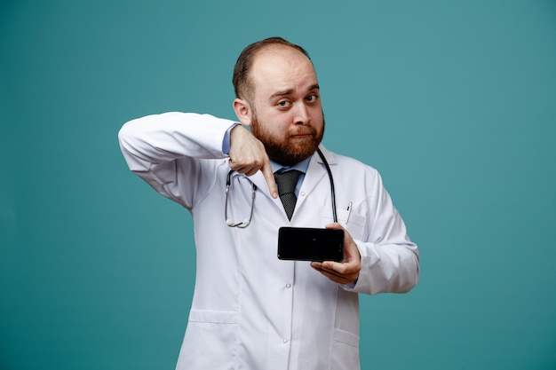 Impresionado joven médico masculino usando abrigo médico y estetoscopio alrededor de su cuello mostrando el teléfono móvil apuntándolo mirando a la cámara aislada en el fondo azul