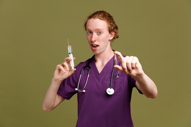 Foto gratuita impresionado joven médico masculino vistiendo uniforme con estetoscopio sosteniendo jeringa con ampolla aislada sobre fondo verde