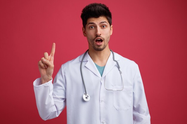 Impresionado joven médico masculino vistiendo uniforme médico y estetoscopio alrededor de su cuello mirando a la cámara apuntando hacia arriba aislado sobre fondo rojo.