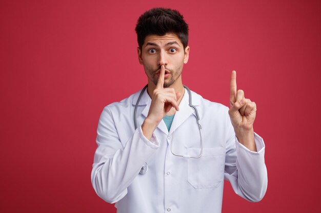 Impresionado joven médico masculino vistiendo uniforme médico y estetoscopio alrededor de su cuello mirando a la cámara apuntando con el dedo hacia arriba aislado sobre fondo rojo.