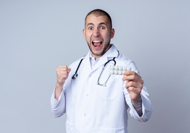 Impresionado joven médico vistiendo una bata médica y un estetoscopio alrededor de su cuello estirando el paquete de tabletas médicas y apretando el puño aislado en blanco