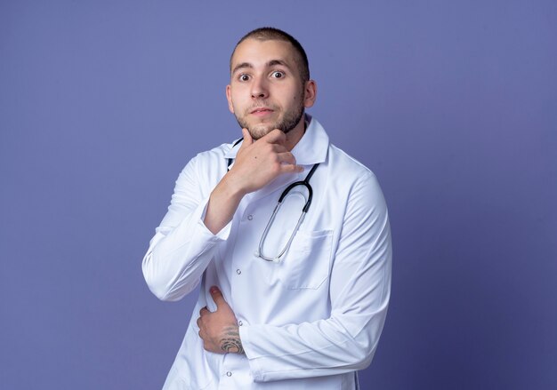 Impresionado joven médico vistiendo una bata médica y un estetoscopio alrededor de su cuello poniendo la mano en el vientre y tocando la barbilla aislada en púrpura