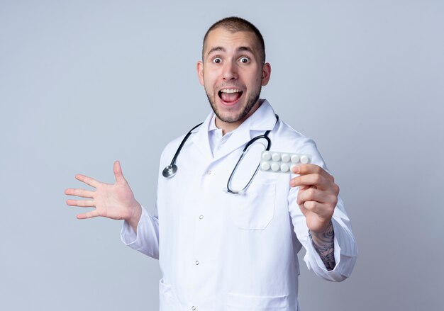 Impresionado joven médico vistiendo bata médica y estetoscopio alrededor de su cuello sosteniendo un paquete de tabletas médicas y mostrando la mano vacía aislada en blanco