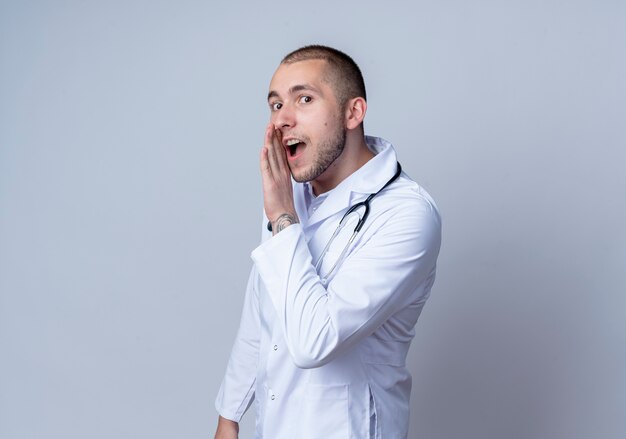 Impresionado joven médico vistiendo bata médica y un estetoscopio alrededor de su cuello susurrando con la mano cerca de la boca aislada en blanco