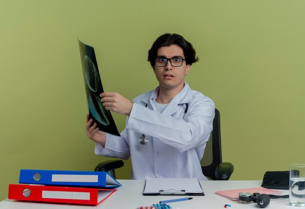 Impresionado joven médico vistiendo bata médica y un estetoscopio con gafas sentado en el escritorio con herramientas médicas con toma de rayos x mirando aislado