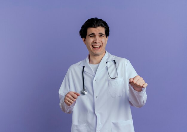 Impresionado joven médico vistiendo bata médica y un estetoscopio mirando al lado manteniendo las manos en el aire aislado
