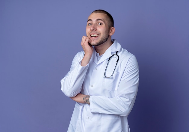 Impresionado joven médico vistiendo bata médica y estetoscopio poniendo las manos en la barbilla y debajo del codo aislado en púrpura