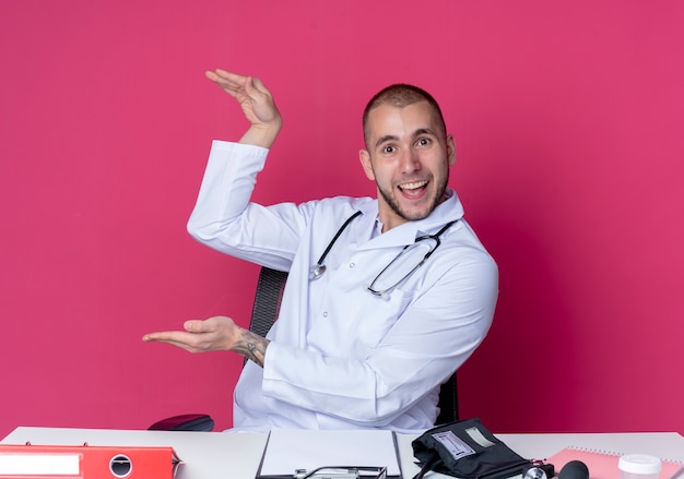 Impresionado joven médico vistiendo bata médica y estetoscopio sentado en el escritorio con herramientas de trabajo que muestran el tamaño con las manos aisladas en rosa