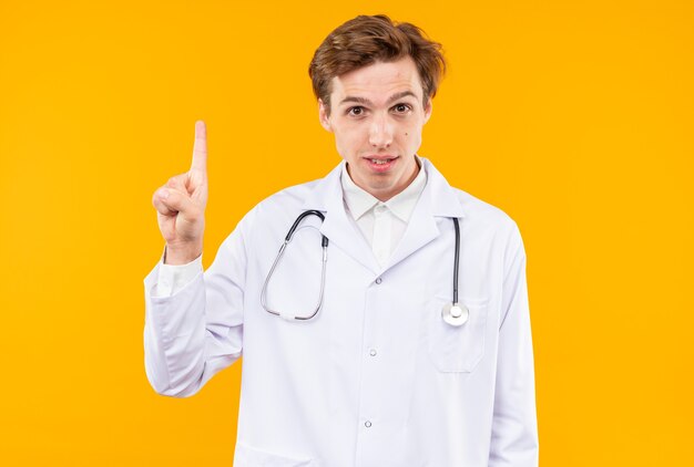 Impresionado joven médico vistiendo bata médica con puntos de estetoscopio en arriba aislado en la pared naranja