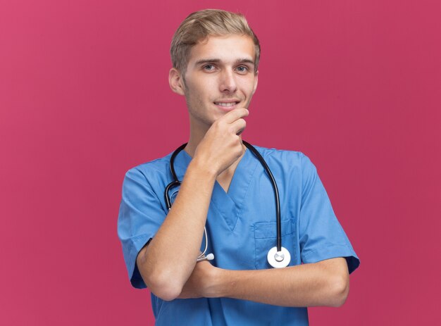 Foto gratuita impresionado joven médico vistiendo uniforme médico con estetoscopio agarró la barbilla aislada en la pared rosa