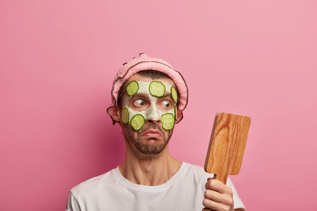 Impresionado joven mira al espejo de madera, aplica mascarilla de arcilla y pepinos, sorprendido de tener muchas arrugas en la tez