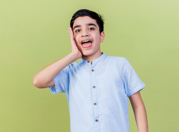 Impresionado joven mirando al frente manteniendo la mano en la cara aislada en la pared verde oliva