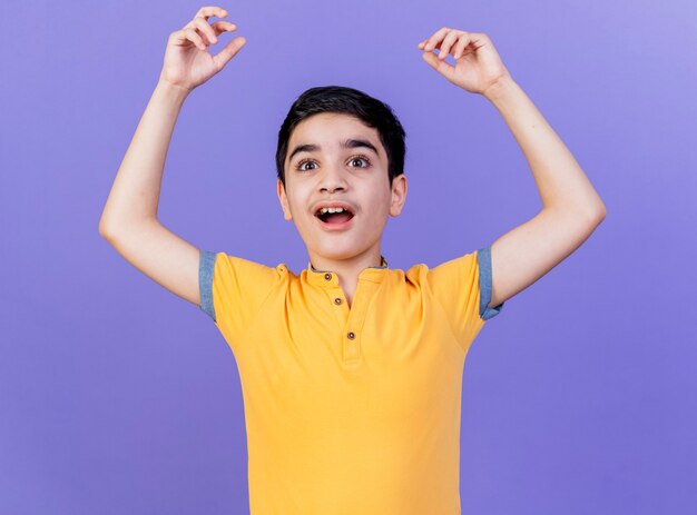 Impresionado joven muchacho caucásico mirando directamente levantando las manos aisladas en la pared púrpura