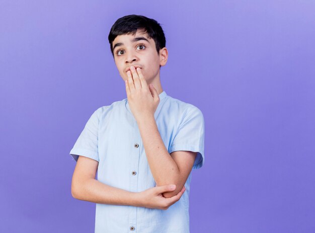 Impresionado joven muchacho caucásico tocando la boca mirando directamente aislado sobre fondo púrpura con espacio de copia