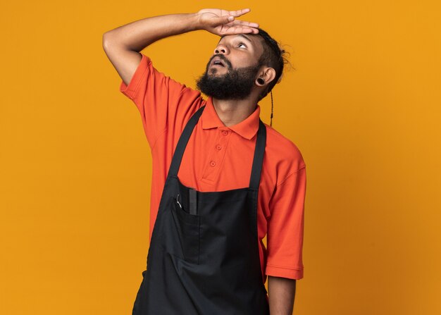 Impresionado joven peluquero afroamericano vistiendo uniforme tocando la frente mirando hacia arriba aislado en la pared naranja con espacio de copia