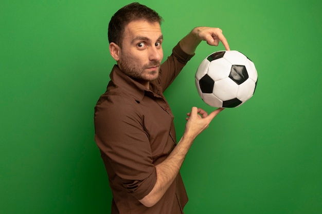 Impresionado joven de pie en la vista de perfil sosteniendo un balón de fútbol mirando al frente aislado en la pared verde