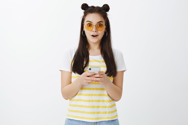 Impresionado joven posando con gafas de sol y teléfono contra la pared blanca