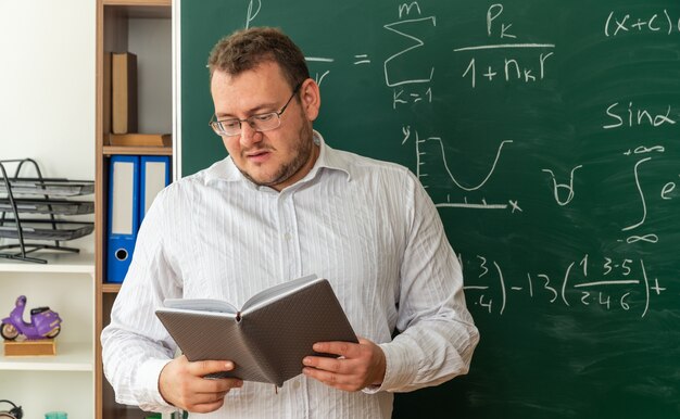 Impresionado joven profesor con gafas de pie delante de la pizarra en el aula leyendo el bloc de notas