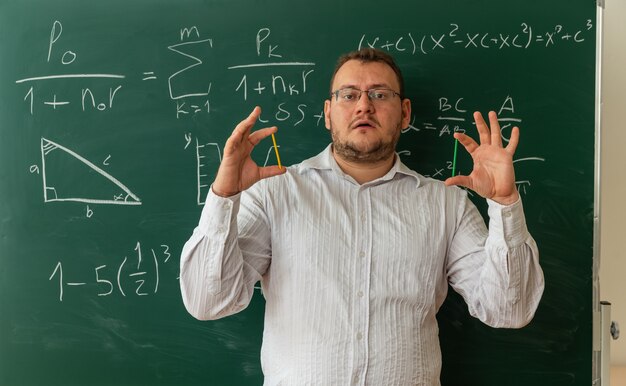 Impresionado joven profesor con gafas de pie delante de la pizarra en el aula mirando al frente mostrando palos de conteo