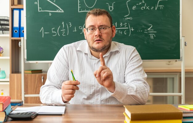 Impresionado joven profesor con gafas sentado en un escritorio con útiles escolares en el aula sosteniendo palos de conteo mirando al frente levantando el dedo