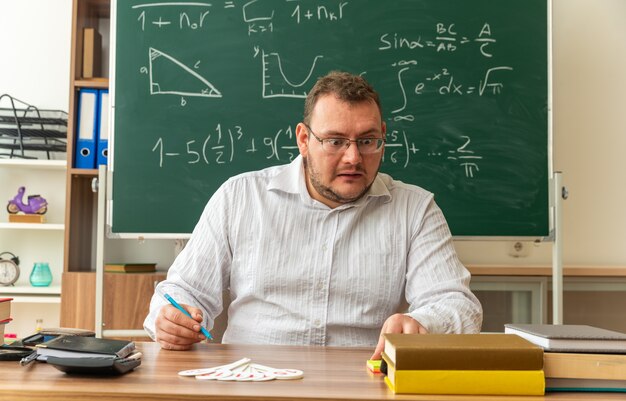 Impresionado joven profesor con gafas sentado en un escritorio con útiles escolares en el aula sosteniendo la pluma tocando y mirando notas de papel
