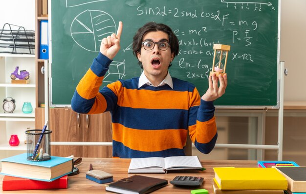 Impresionado joven profesor de geometría con gafas sentado en un escritorio con útiles escolares en el aula sosteniendo un reloj de arena mirando al frente apuntando hacia arriba